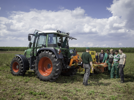 Ein Traktor steht auf einer Wiese, daneben junge Auszubildende. Dahinter ein Feld und blauer Himmel.
